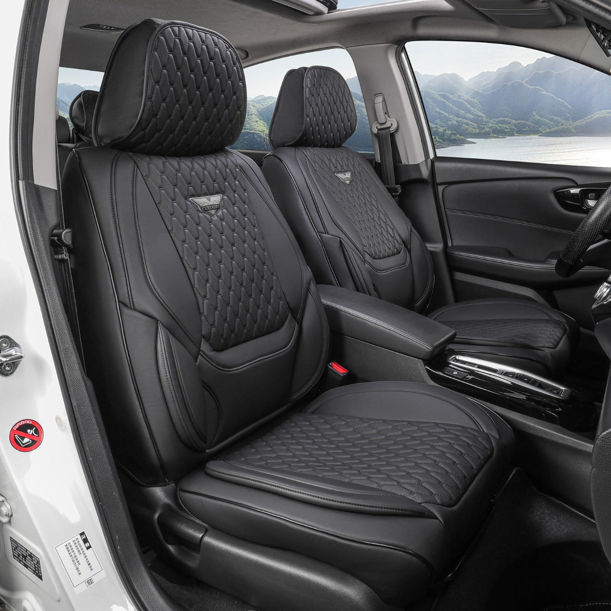 Black car seat covers installed on the front seats of a vehicle, featuring quilted diamond stitching and a small wing logo on the backrests, shown from the open driver's side door with a scenic mountain lake view outside.