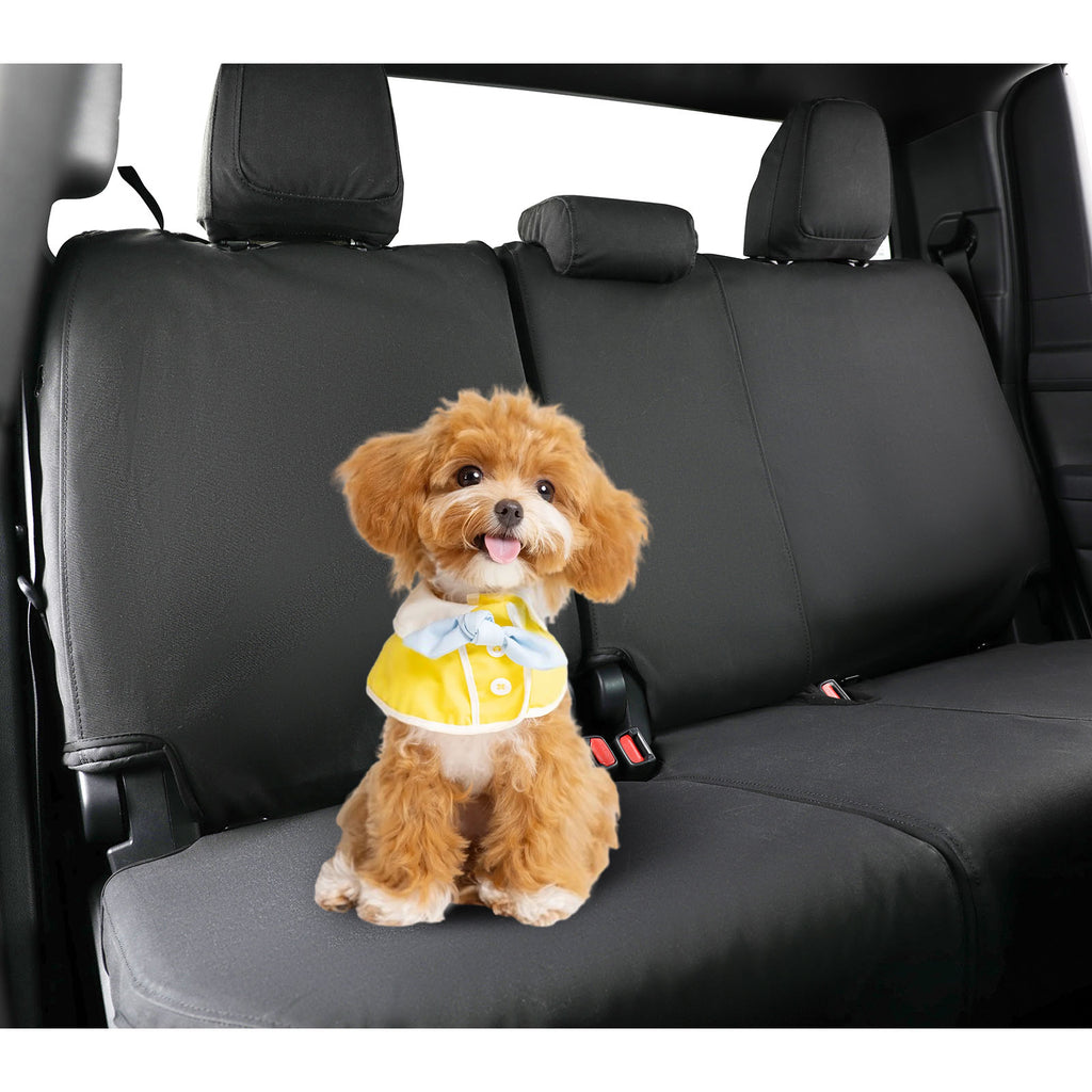 A small fluffy brown dog wearing a yellow vest with a blue bow sits on a black pet car seat inside a vehicle. The image highlights the black car seat cover and harness system.