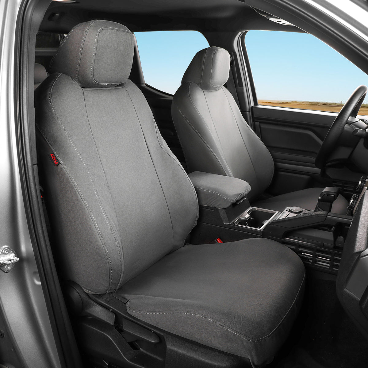 A pair of gray car seat covers installed on the front seats of a vehicle interior, shown with the driver-side door open and a landscape visible through the windows.