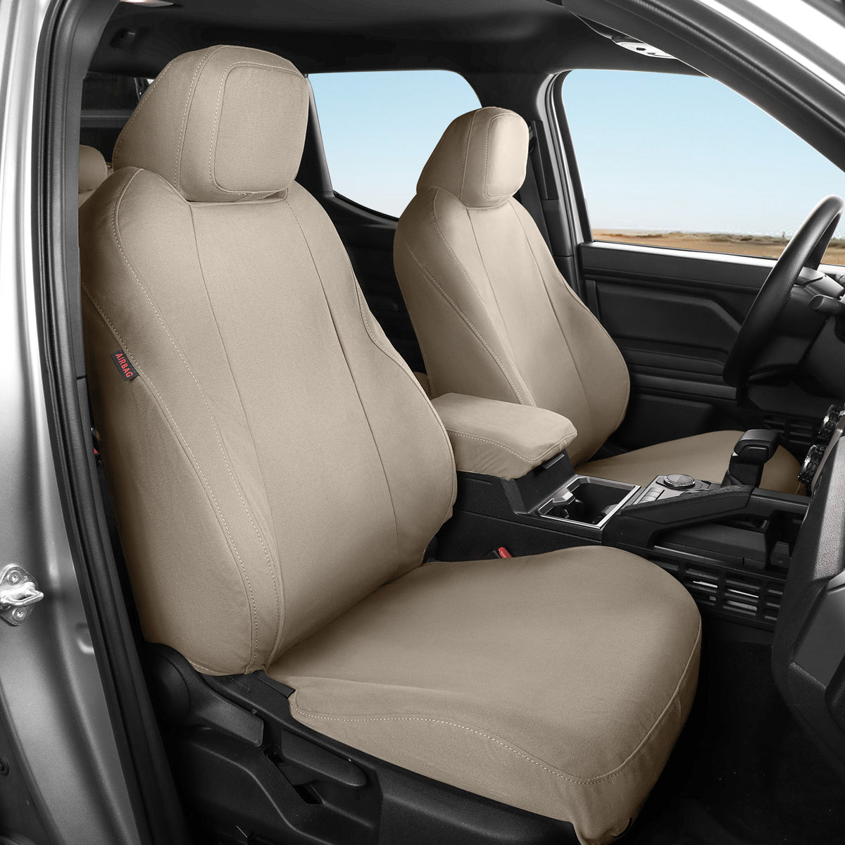 Tan car seat covers installed on the front seats of a vehicle interior, shown from the open driver-side door with the dashboard, center console, and windows visible.