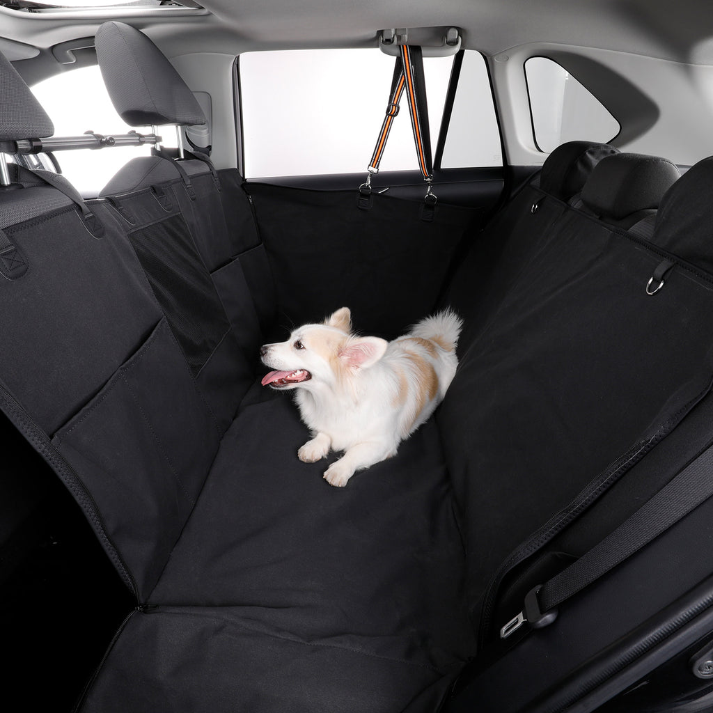 A small white and tan dog lies on a black car seat cover installed across the rear bench seats inside a vehicle.