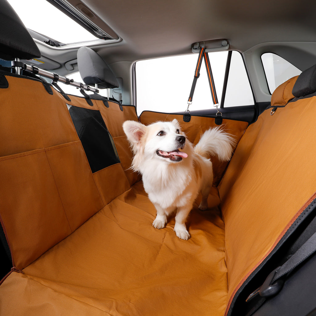 A small fluffy white dog stands on an orange pet car seat cover inside a car trunk. The cover is shown stretched across the back seat area, with black side panels and a harness attachment visible.