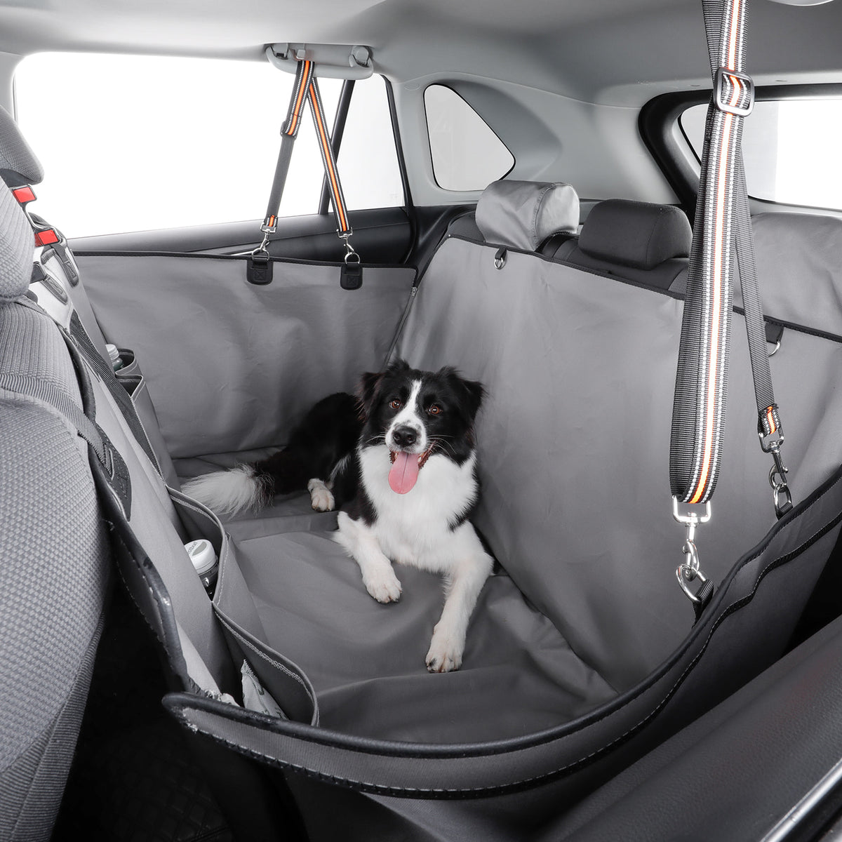 A black and white dog lies on a gray car seat cover installed in the back of a car. The cover includes side panels and hanging straps, and the dog is positioned in the center of the vehicle interior.