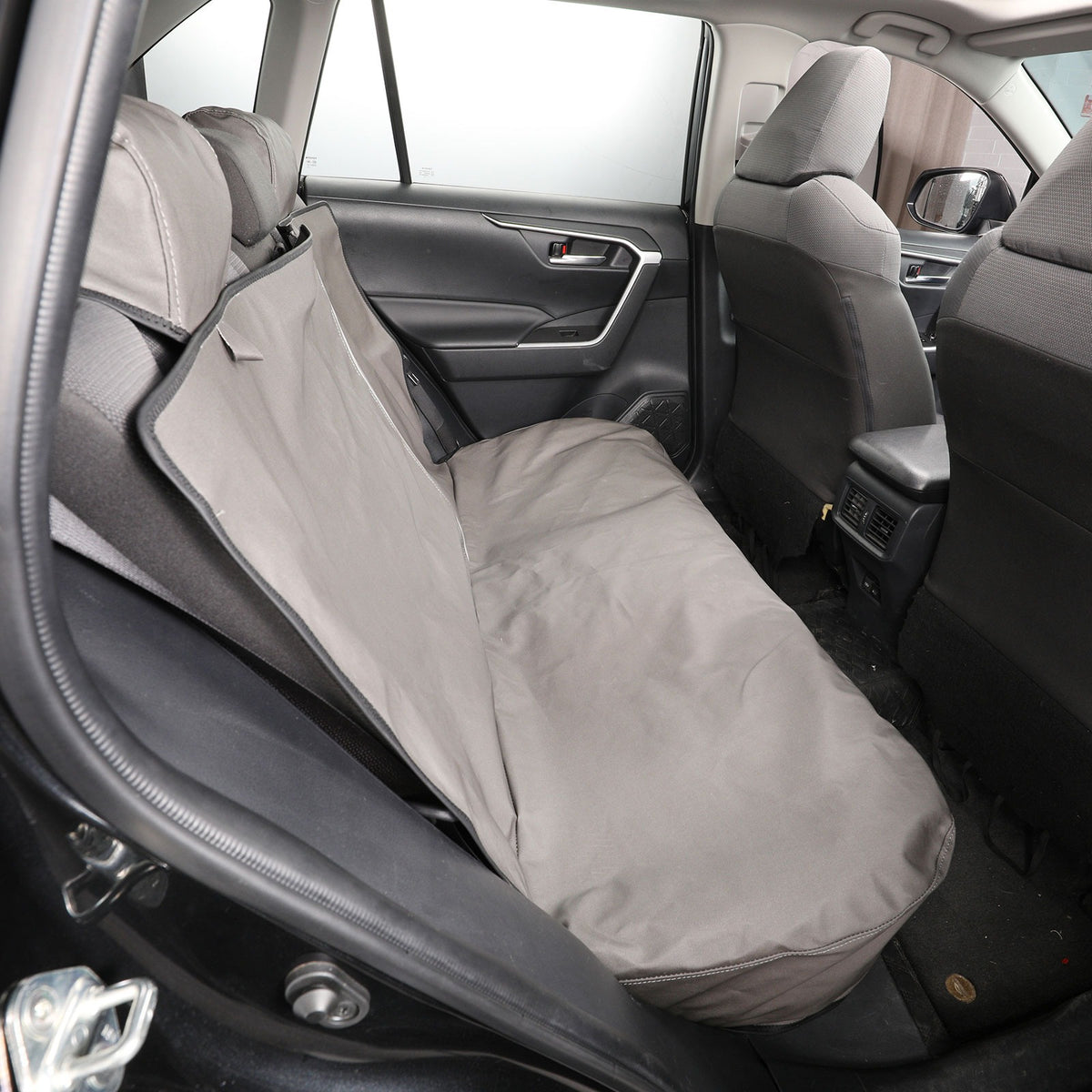 A light gray car seat cover installed on the rear bench seat of a vehicle interior, shown from the open rear door.