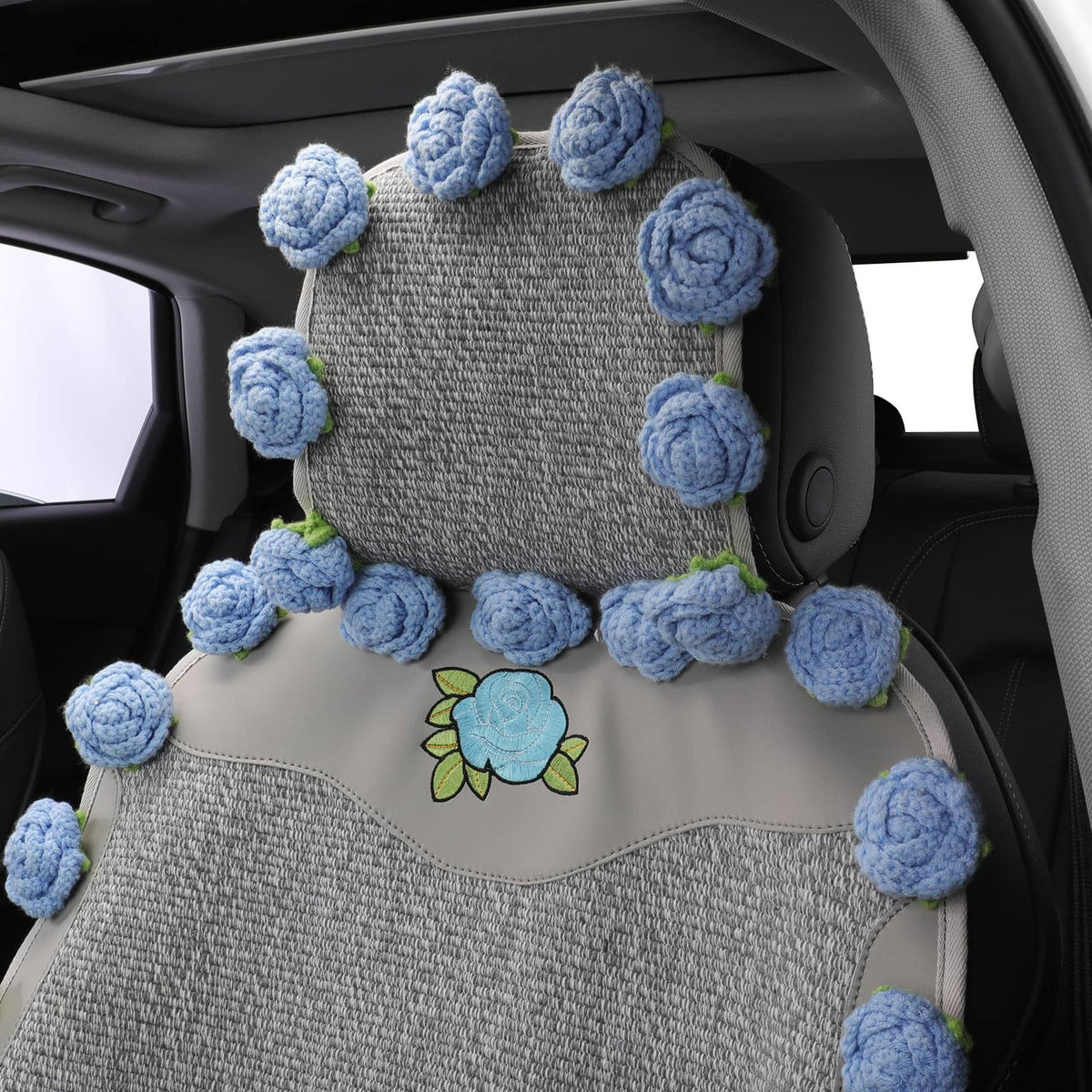 A car seat cover set with a textured gray fabric base and decorative light blue crocheted roses around the headrest and backrest, plus a blue rose and green leaf motif on the seat cushion, shown installed inside a vehicle.