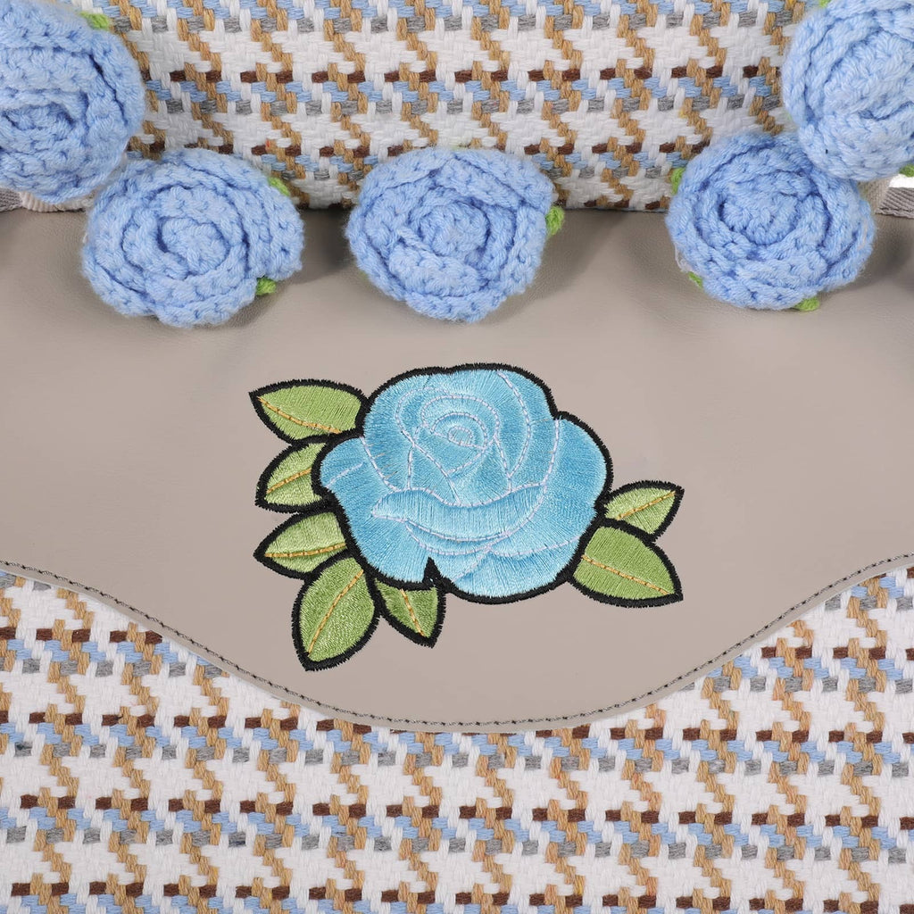A close-up of a beige leather or faux-leather patch featuring an embroidered light blue rose with green leaves, bordered by a wavy stitched edge. The background shows a woven textile in beige, brown, and light blue, with several matching light blue crocheted rose embellishments arranged along the top.