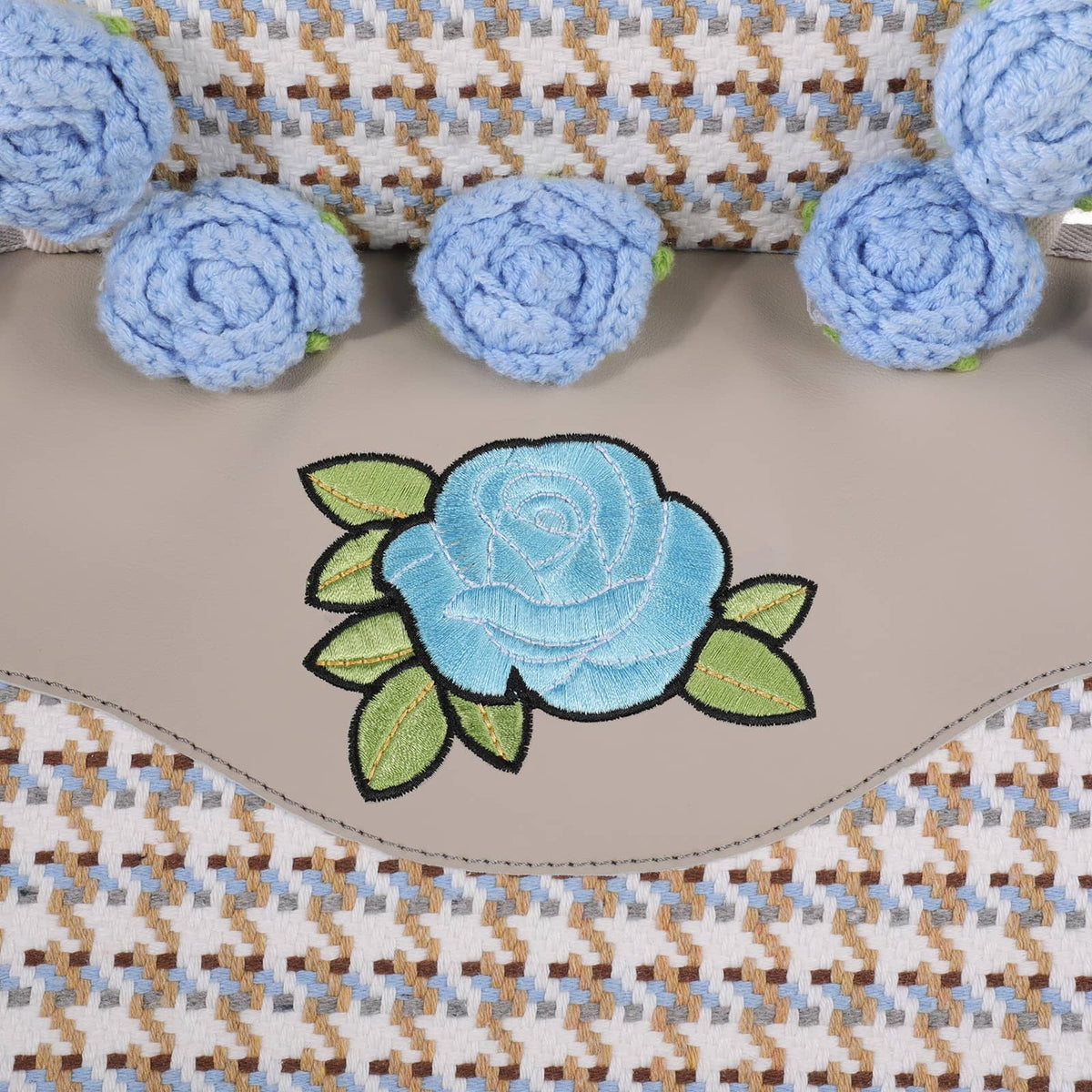 A close-up of a beige leather or faux-leather patch featuring an embroidered light blue rose with green leaves, bordered by a wavy stitched edge. The background shows a woven textile in beige, brown, and light blue, with several matching light blue crocheted rose embellishments arranged along the top.